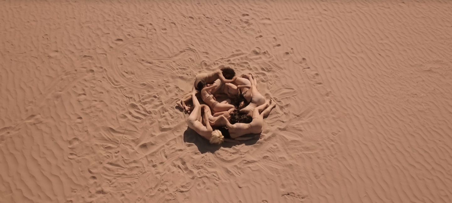 A group  of six people are laying down on a sand surface with their bodies intertwined. They are dressed in underwear matching their skintone, making them appear as nude.