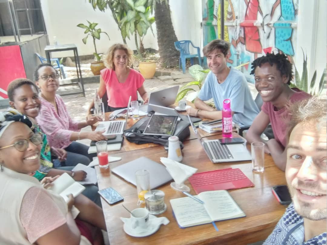 seven people sitting around a table outside in the sunshine. They are smiling at the camera. On the table there are laptops and notepads.